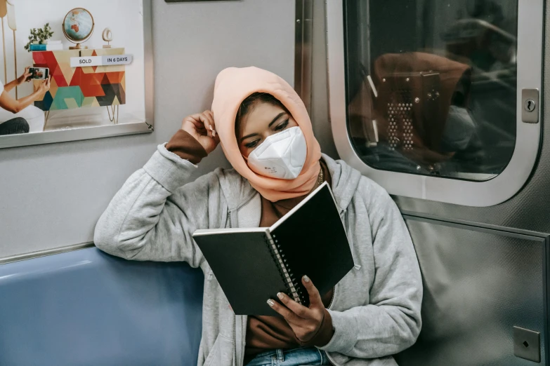 a woman sitting on a train reading a book, pexels contest winner, hurufiyya, surgical mask covering mouth, riyahd cassiem, holding notebook, without makeup