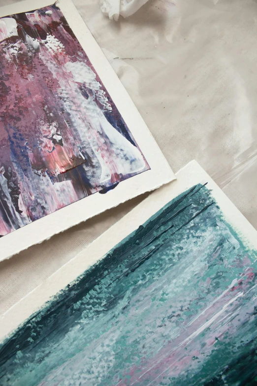 a couple of paintings sitting on top of a table, a watercolor painting, inspired by Yanjun Cheng, trending on unsplash, screen print texture, close - up photograph, textured paper, mauve and cyan