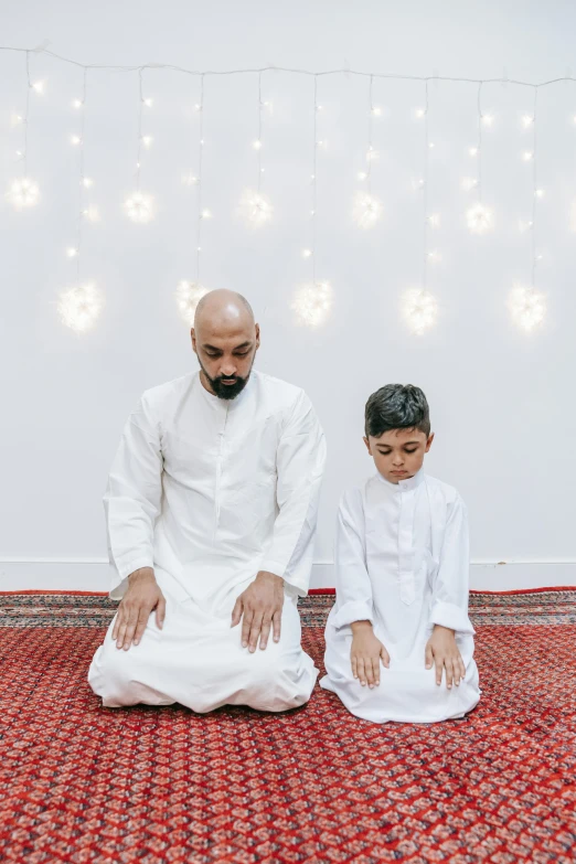 a man and a boy are sitting on the floor, hurufiyya, doing a prayer, lights on, forward facing pose, fatherly