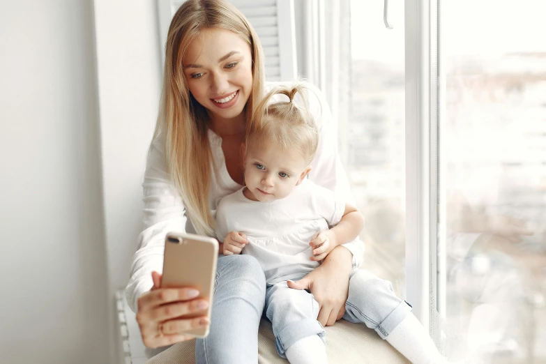 a woman holding a baby and looking at a cell phone, a picture, pexels contest winner, a blond, near the window, avatar image, looking towards camera