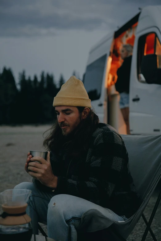 a man sitting in a chair next to a campfire, trending on unsplash, renaissance, mid long hair, snake van, wearing a beanie, coffee cup