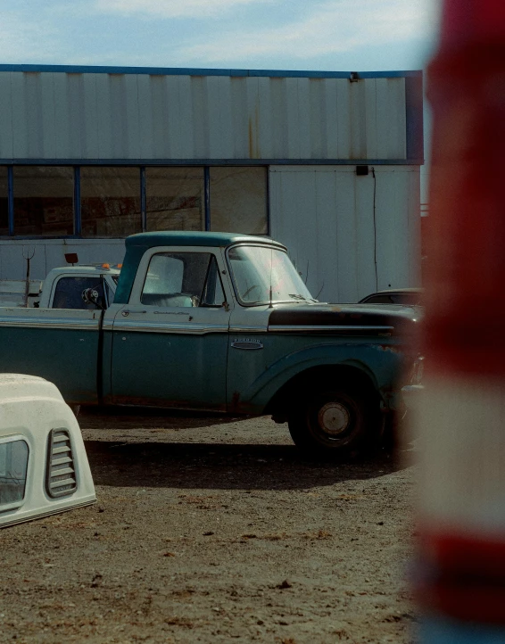 a blue truck parked in front of a building, an album cover, unsplash, photorealism, movie footage, scrapyard, faded color film, 4 k film still