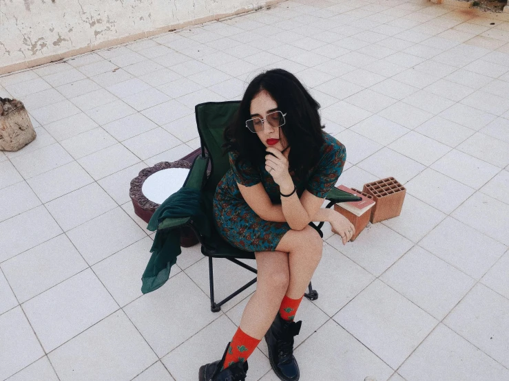 a woman sitting in a chair talking on a cell phone, an album cover, inspired by Elsa Bleda, pexels contest winner, graffiti, young middle eastern woman, girl wearing round glasses, she wears a dark green dress, ootd