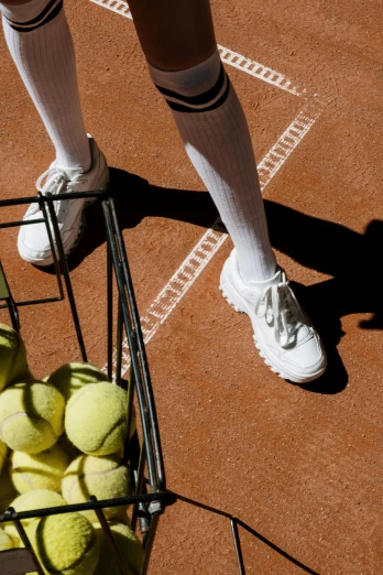 a person standing on a tennis court with a basket full of tennis balls, inspired by Hans Mertens, white uniform, shoes, terracotta, up close
