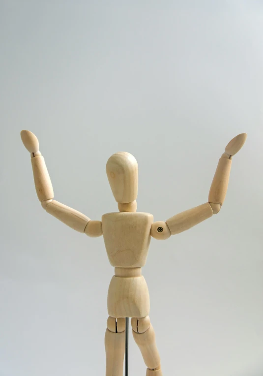 a wooden mannequin holding his arms in the air, unsplash, visual art, plain background, five points of articulation, back turned, toy photography