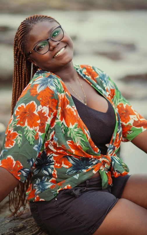 a woman sitting on a rock next to a body of water, by Chinwe Chukwuogo-Roy, trending on pexels, with hawaiian shirt, madly grinning, girl with glasses, brown:-2