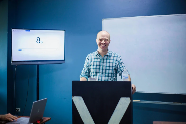 a man standing at a podium in front of a projector screen, unsplash, academic art, vitalik buterin, eight eight eight, karl pilkington, in a classroom