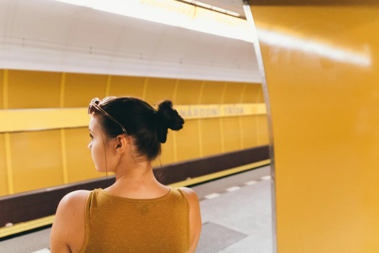 a woman standing in a train station waiting for a train, by Emma Andijewska, pexels contest winner, wearing yellow croptop, hair styled in a bun, a round minimalist behind, underground metro