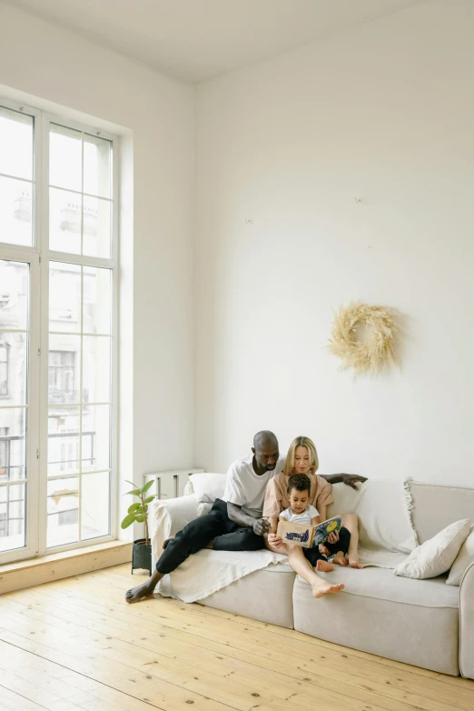 a family sitting on a couch in a living room, by Andries Stock, pexels contest winner, minimalism, city apartment cozy calm, light beige pillows, dwell, natural window lighting