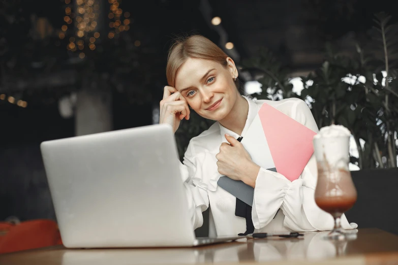 a woman sitting at a table with a laptop, trending on pexels, wearing lab coat and a blouse, holding books, smug expression, thumbnail