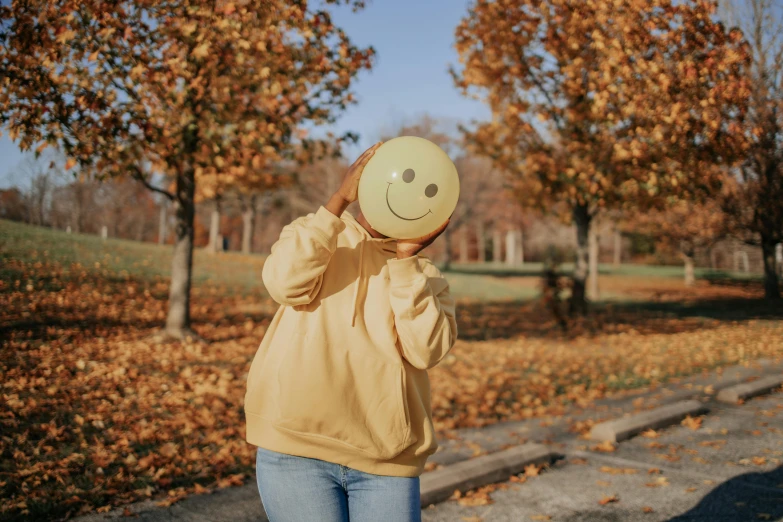 a woman holding a smiley face in front of her face, a photo, by Attila Meszlenyi, pexels contest winner, 🍂 cute, wearing sweatshirt, beautiful day, holding a gold bag