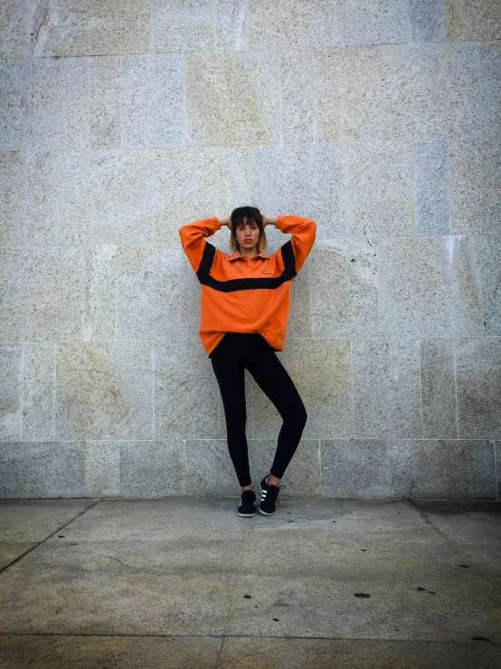 a woman leaning against a wall with her hands on her head, an album cover, by Robbie Trevino, trending on unsplash, long orange sweatshirt, 8 0 s sport clothing, full body pose, orange and black