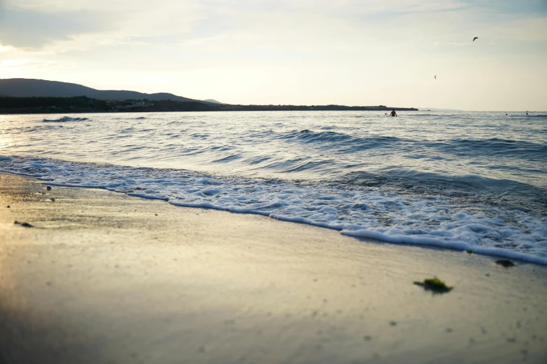 a person flying a kite on top of a sandy beach, body of water, in the evening, from the distance, wavy water