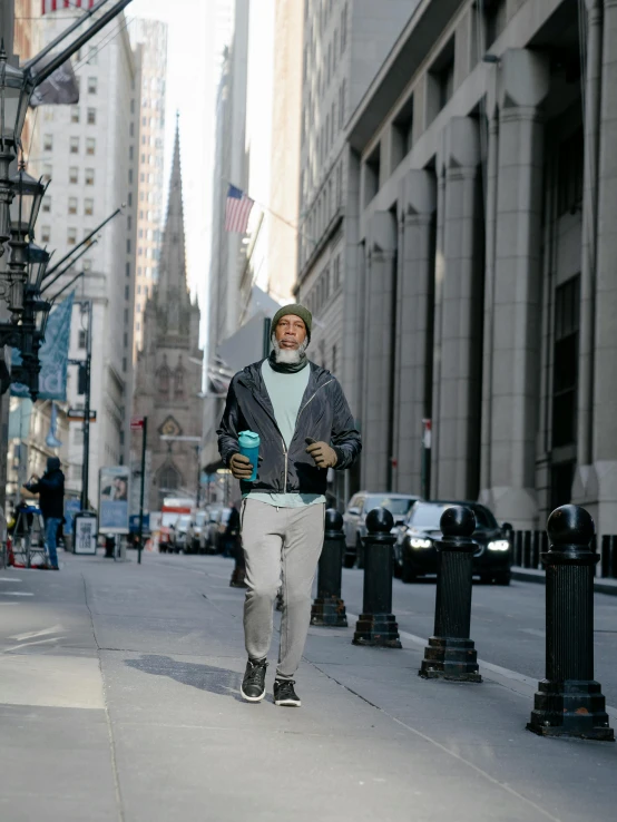 a man walking down a city street next to tall buildings, wearing teal beanie, grey trimmed beard, full body potrait holding bottle, ( ( theatrical ) )