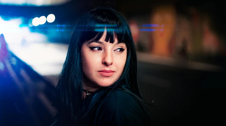 a woman standing in the middle of a street at night, an album cover, by Julia Pishtar, pexels contest winner, photorealism, long black hair with bangs, police lights shine on her face, headshot profile picture, blue color grading