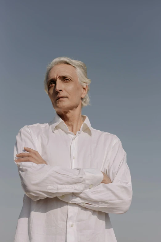 a man in a white shirt standing with his arms crossed, an album cover, inspired by John Cale, unsplash, photorealism, silver haired, androgynous person, portrait of bertrand russell, profile pic