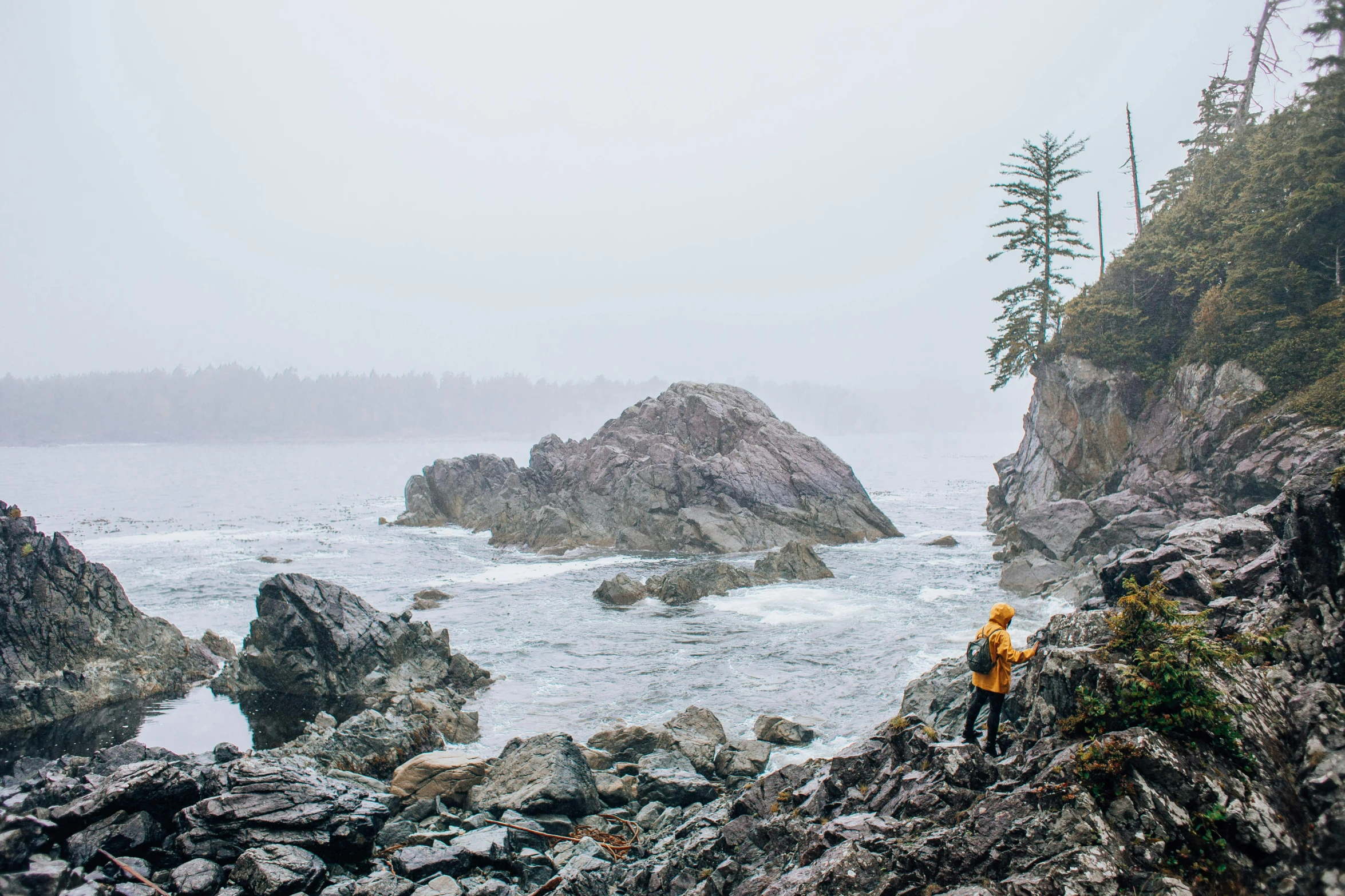 a person sitting on a rock near a body of water, a photo, pacific northwest coast, grey skies rain, conde nast traveler photo, wearing adventuring gear