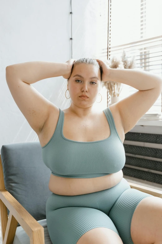 a woman sitting in a chair next to a window, a colorized photo, trending on pexels, her belly is fat and round, sport bra and shirt, teal skin, frown fashion model