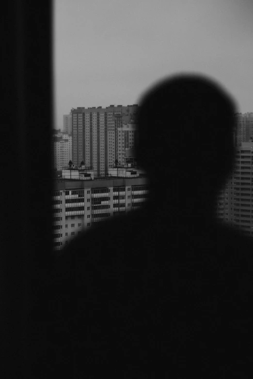 a black and white photo of a person looking out a window, inspired by Cheng Jiasui, city background in silhouette, self - portrait!!!!, hello darkness my old friend, disolate :: a long shot