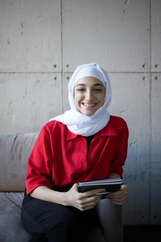 a woman sitting on a couch holding a tablet computer, an album cover, inspired by Maryam Hashemi, shutterstock contest winner, hurufiyya, red shirt, white hijab, smiling young woman, mit technology review