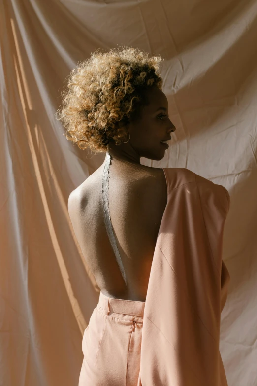 a woman standing in front of a white backdrop, trending on pexels, renaissance, short blonde afro, open back dress, pink golden hour, jacket over bare torso