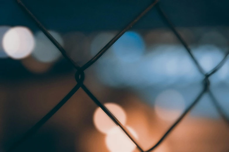 a close up of a chain link fence with lights in the background, inspired by Elsa Bleda, trending on pexels, visual art, background image, unfocused, night photo, alessio albi