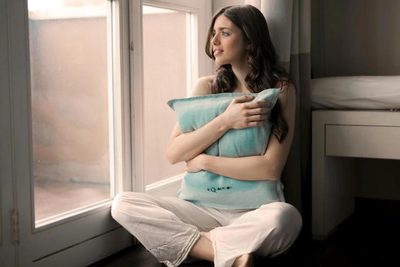 a woman sitting in front of a window holding a pillow, aquamarine windows, sleepy expression, ivory, medium-shot