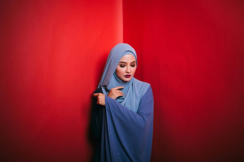 a woman in a blue hijab leaning against a red wall, pexels contest winner, hurufiyya, grey and blue theme, “diamonds, red clothes, a young asian woman