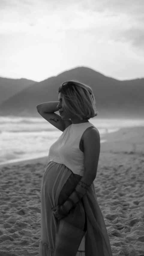 a woman standing on top of a beach next to the ocean, a black and white photo, by Emerson Silva, pexels contest winner, happening, pregnant belly, 3 5 year brazilian mother, profile pic, concert