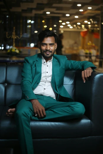 a man in a green suit sitting on a black couch, inspired by Saurabh Jethani, pexels contest winner, confident pose, white and teal garment, lgbtq, evening time