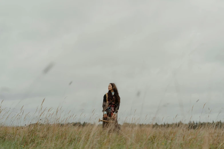 a woman standing in a field of tall grass, an album cover, unsplash, grey skies, portrait image, autumn season, video still