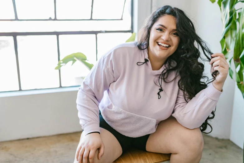 a woman sitting on top of a wooden chair, inspired by Ion Andreescu, trending on pexels, renaissance, wearing a pastel pink hoodie, her belly is fat and round, dark short curly hair smiling, plus size woman