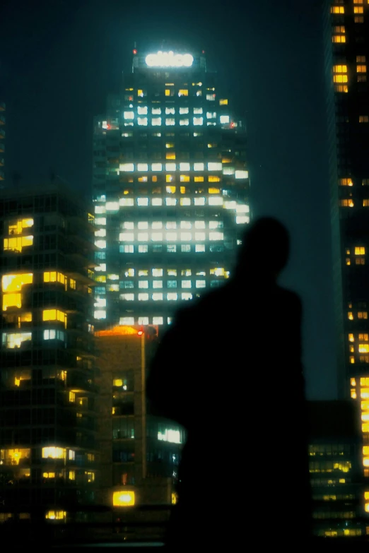 a man standing in front of a tall building at night, inspired by roger deakins, neo-figurative, silhouetted, canary wharf, viewed from a distance, out of focus