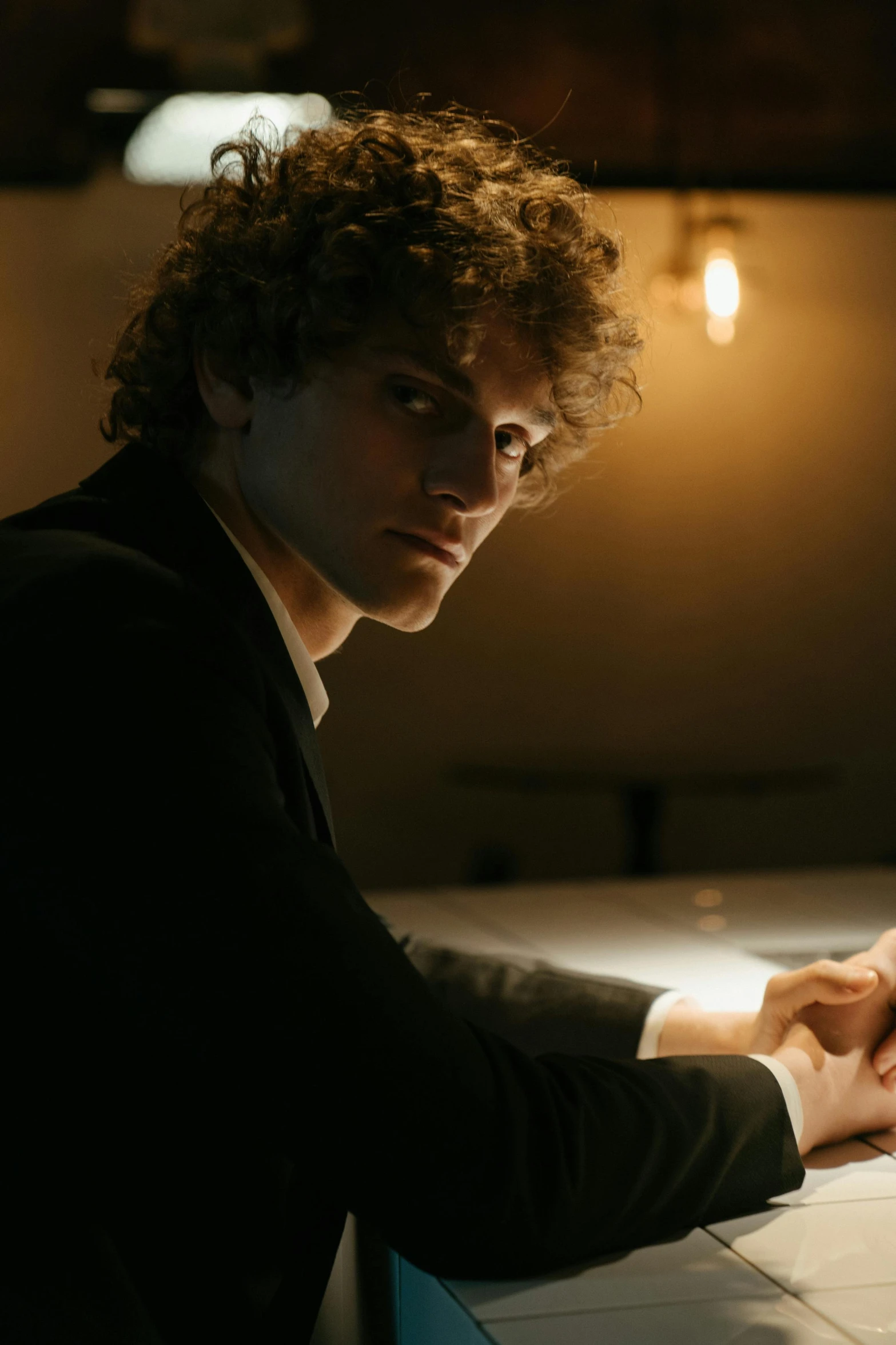 a man sitting at a table in front of a laptop computer, an album cover, inspired by Nan Goldin, trending on pexels, digital art, curly haired, portrait of a man in a suit, chiaroscuro soft lighting, ( ( theatrical ) )