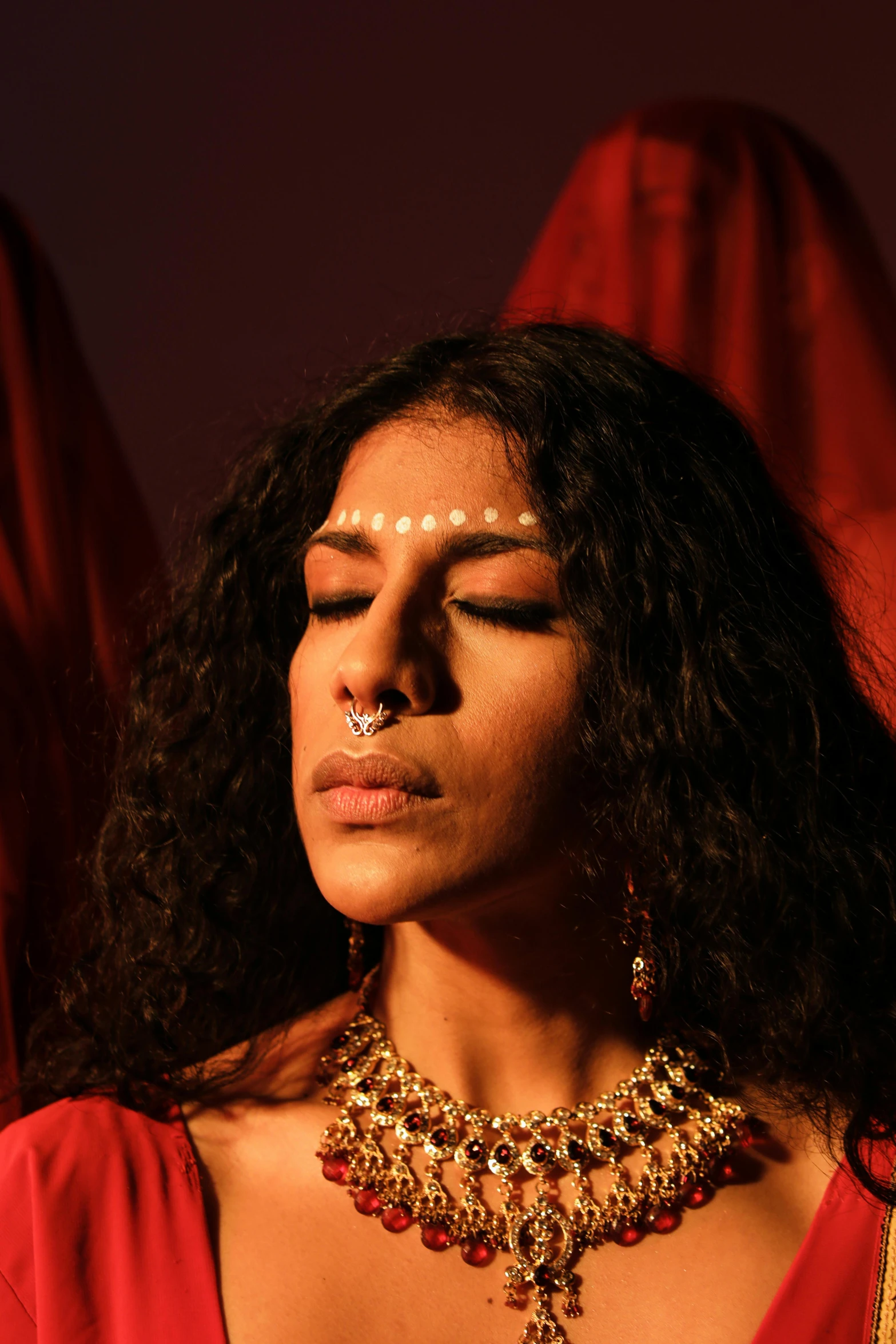 a woman in a red dress with her eyes closed, by Sunil Das, huge earrings and queer make up, hindu stages of meditation, transgender, beatifully lit