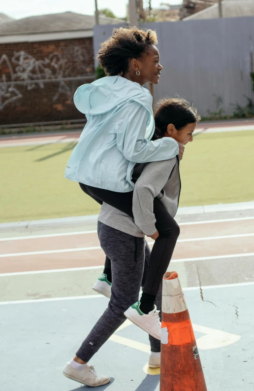 a woman carrying a child across a tennis court, by Jessie Algie, pexels contest winner, happening, wearing track and field suit, head down, stacked image, streetwear