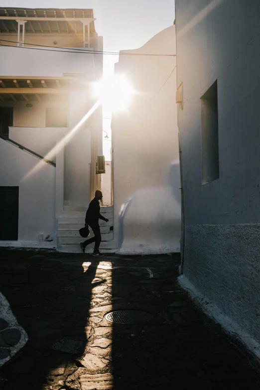 a person walking through a narrow alley way, by Alexis Grimou, light and space, sun at dawn, white building, buildings covered in black tar, greece