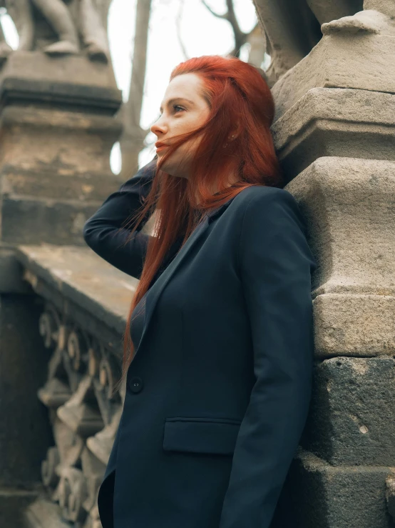 a woman with red hair leaning against a stone wall, inspired by Elsa Bleda, pexels contest winner, wearing dark blue suit, profile image, wearing wool suit, street photo