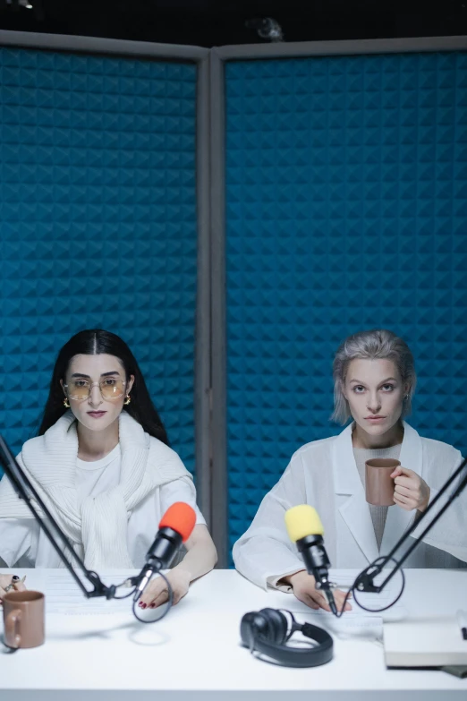 two women sitting at a table with microphones in front of them, inspired by Elsa Bleda, wearing futuristic white suit, looking confused, two hovering twin nuns, grimes