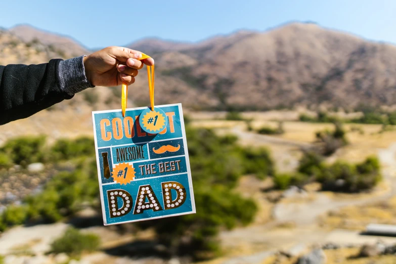 a close up of a person holding a sign, pexels contest winner, dad bod, gold and teal color scheme, mountains in the background, gifts