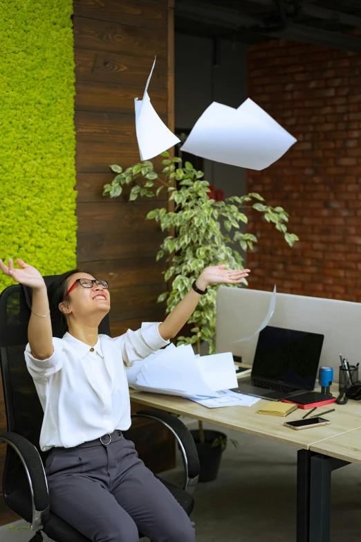 a woman sitting in a chair throwing papers into the air, happening, unhappy, highly upvoted, on a desk, next to a plant