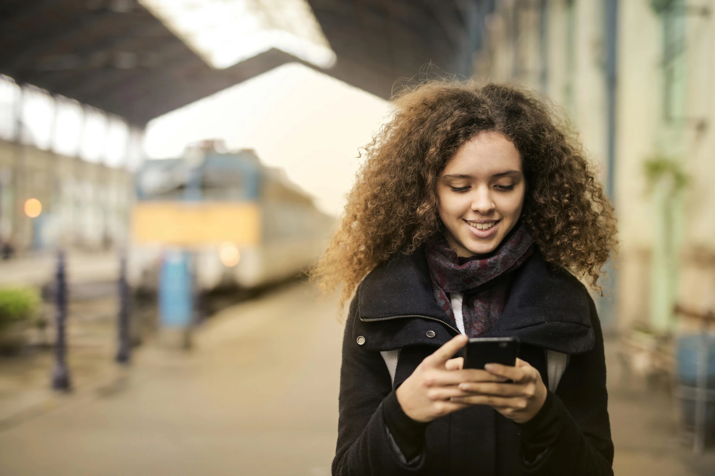 a woman standing in a train station looking at her cell phone, trending on pexels, happening, square, curly haired, teenager, australian