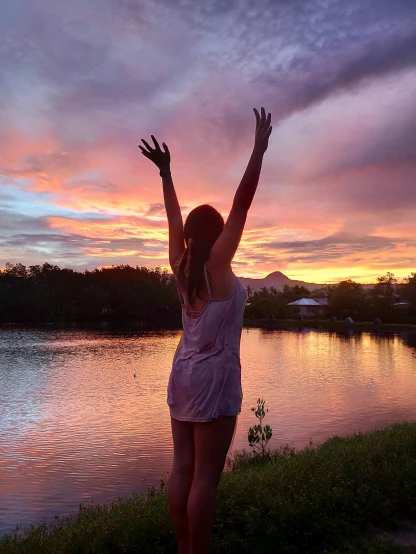 a woman standing next to a body of water at sunset, waving and smiling, erin moriarty, instagram post, arms raised