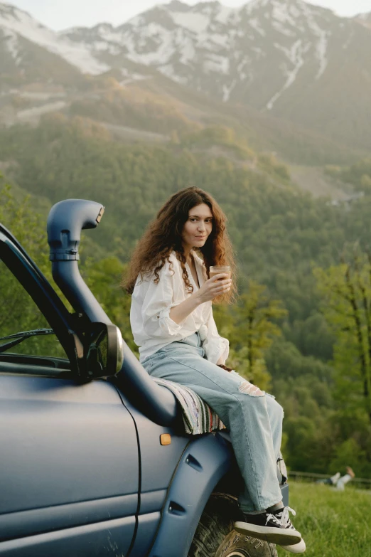 a woman sitting on the hood of a car, trending on pexels, renaissance, holding a bottle of arak, mountainside, long curly brown hair, cottagecore hippie