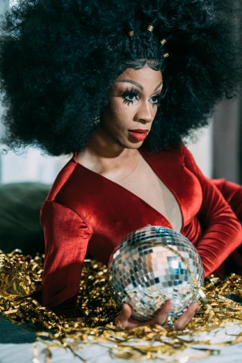 a woman in a red dress holding a disco ball, an album cover, trending on pexels, kitsch movement, afro hair, dressed in velvet, proud looking away, drag