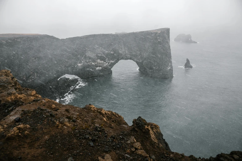 a rock formation in the middle of a body of water, under a gray foggy sky, arch, iceland photography, far view