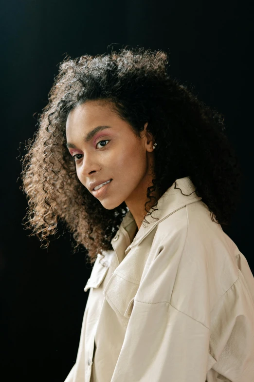 a woman standing in front of a black background, inspired by Afewerk Tekle, trending on pexels, renaissance, wearing a long beige trench coat, a black man with long curly hair, brown skin. light makeup, photo of young woman