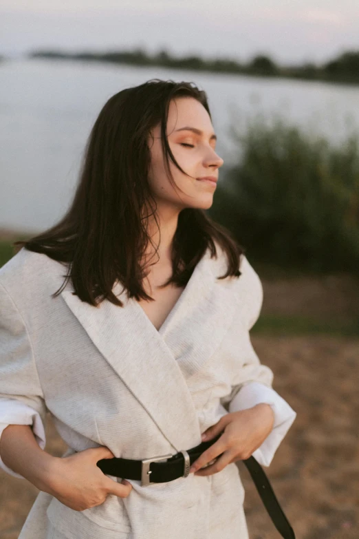 a woman standing in front of a body of water, an album cover, by Attila Meszlenyi, trending on pexels, renaissance, wearing a long beige trench coat, thick collar, summer evening, brunette woman