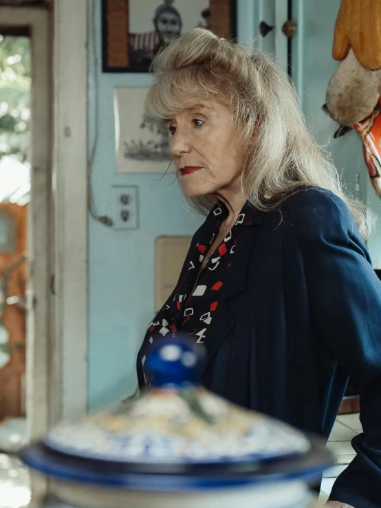 a woman standing in a kitchen next to a counter, a portrait, by Penelope Beaton, indie film, an elderly, profile image, wearing blue jacket