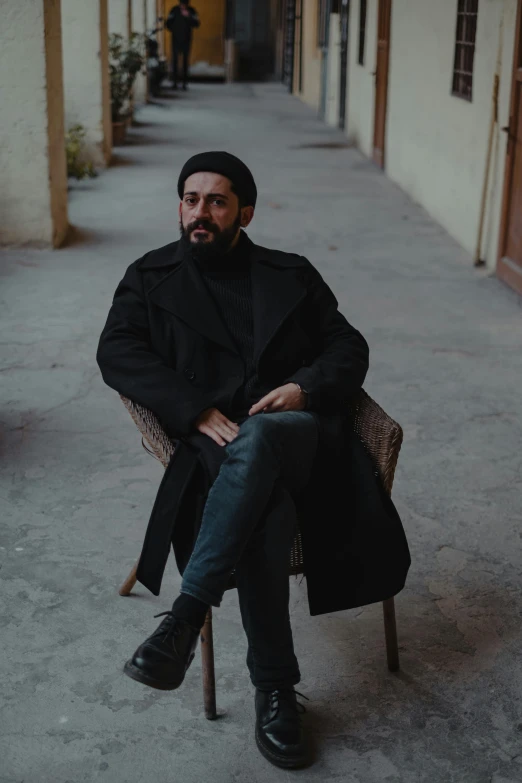 a man sitting on a chair in a hallway, an album cover, inspired by Nabil Kanso, pexels contest winner, with a full black beard, worn black coat, reza afshar, on a street